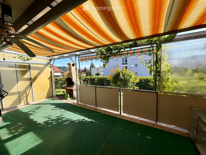 Covered balcony with striped awning, green artificial turf, glass walls, and outdoor furniture.