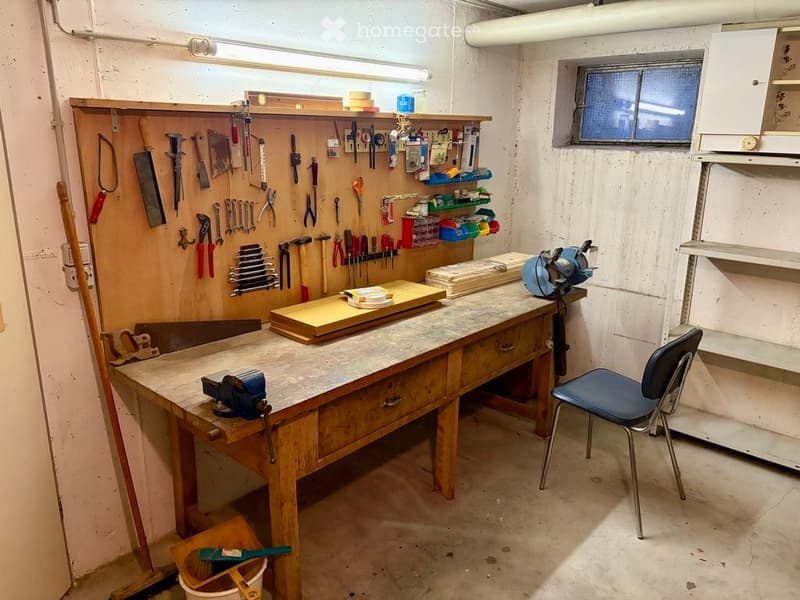 A well-organized workshop with a large wooden workbench, various tools hanging on the wall, a blue chair, and shelves holding additional supplies.