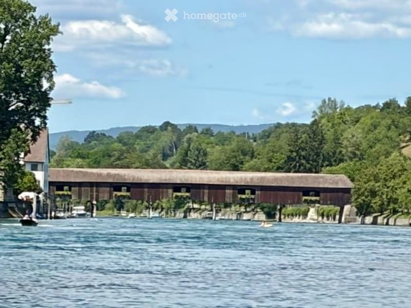 Scenic view of a covered wooden bridge spanning a calm river, surrounded by lush green trees and distant hills under a clear blue sky.