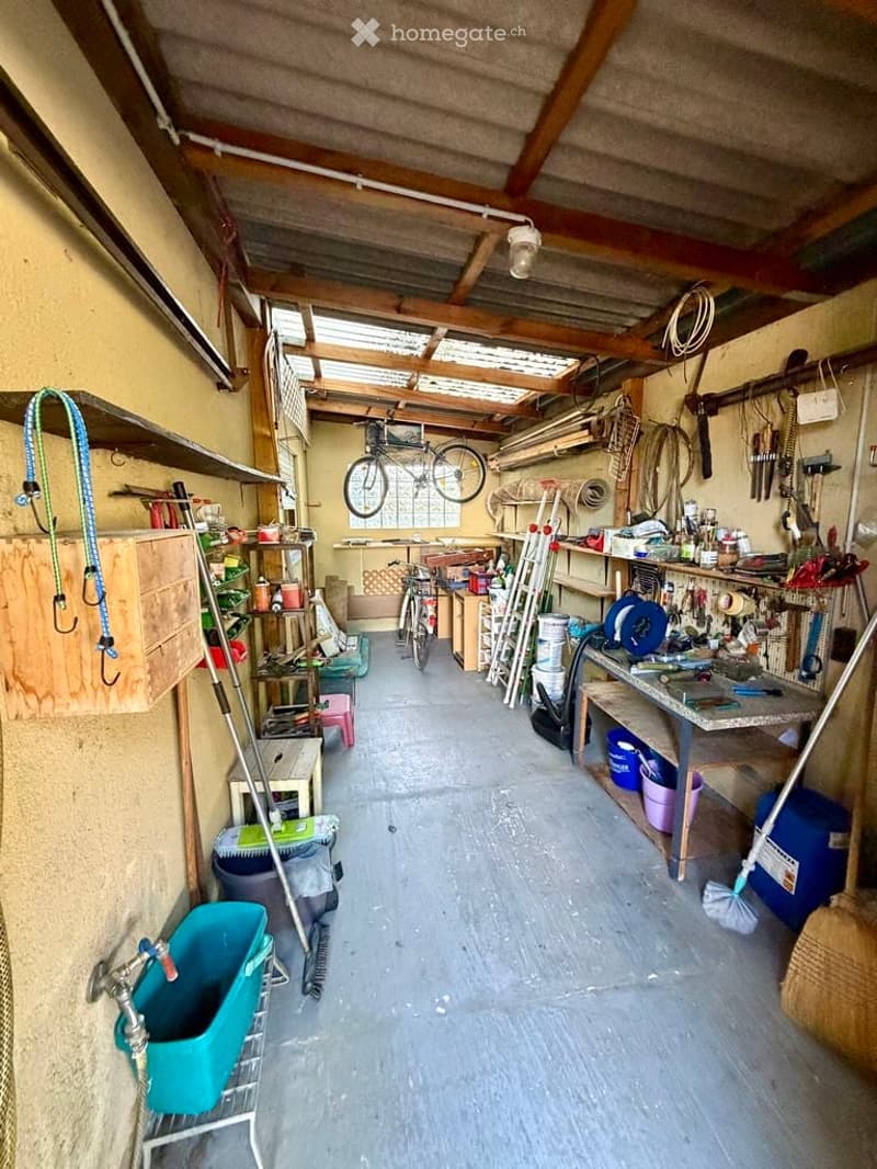 The image shows a well-organized garage with a concrete floor and wooden ceiling beams. Various tools and equipment are neatly stored on shelves and workbenches. A bicycle is hanging from the ceiling, and there are ladders, buckets, and storage boxes visi