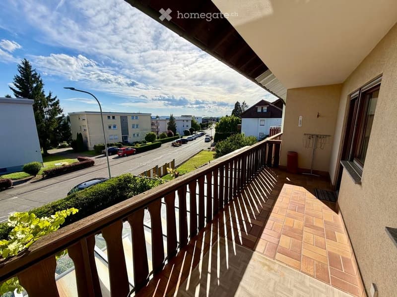 Balcony with wooden railing, tiled floor, overlooking a residential street with parked cars and greenery.
