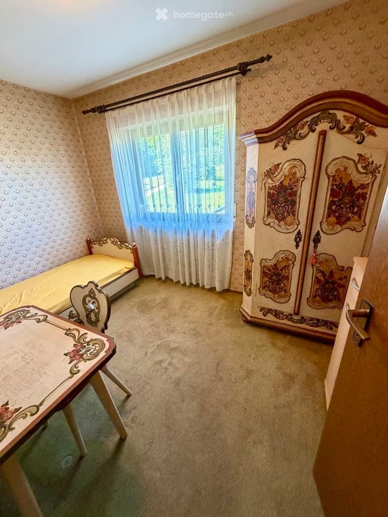 A small, furnished room with a single bed, a small table, a chair, and an ornate wardrobe. The room features patterned wallpaper and a window with sheer curtains.