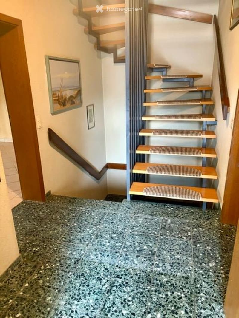 Spacious hallway with a modern staircase featuring wooden steps and metal railings, carpeted treads, and a framed picture on the wall.