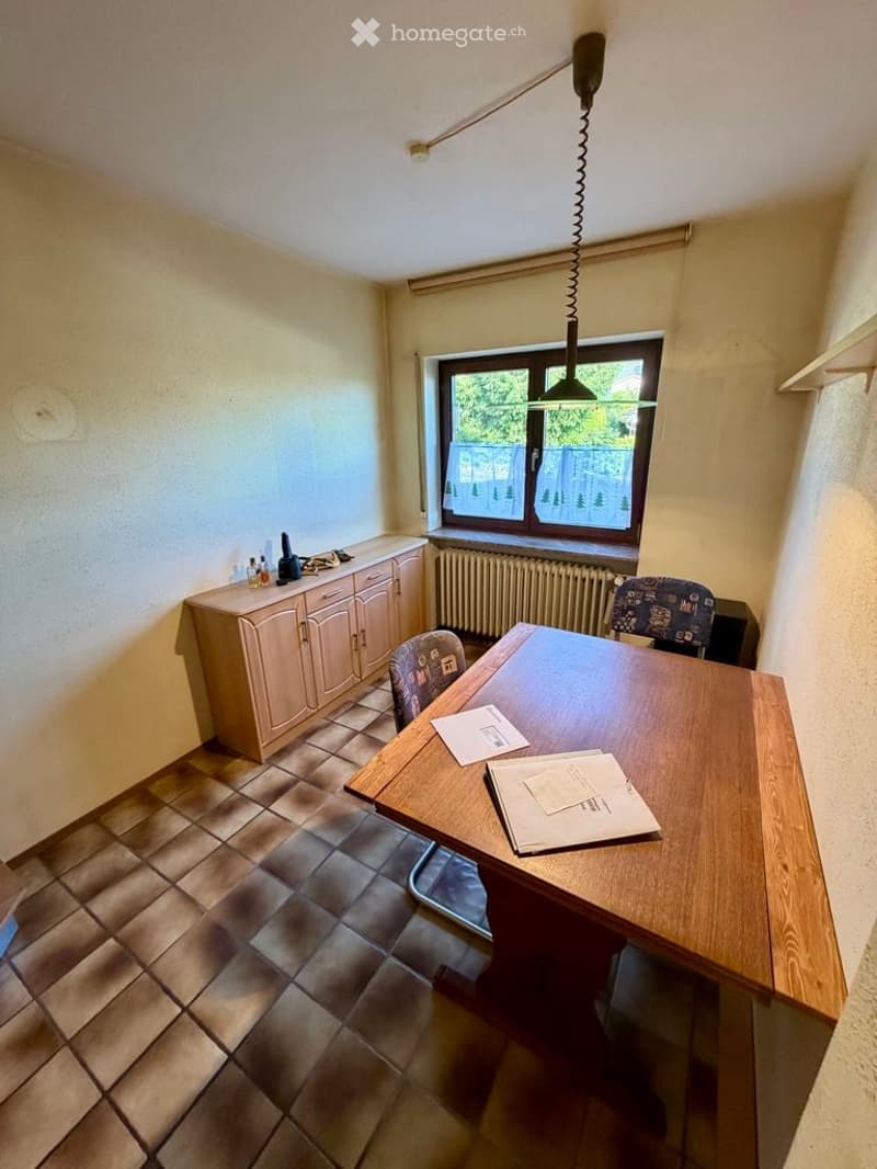 A small, well-lit room with a wooden desk, two chairs, a cabinet, and a radiator under a window. The room has tiled flooring and a hanging light fixture.
