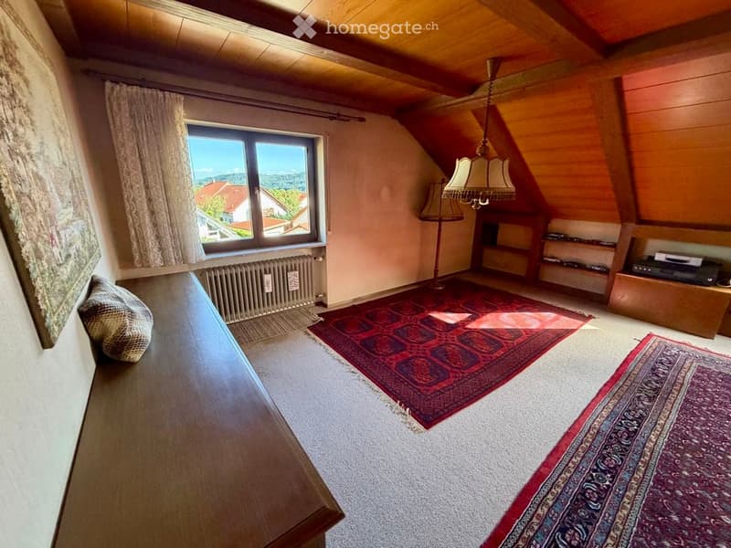Cozy attic room with wooden beams, large window offering a scenic view, a wooden table with a chair, a floor lamp, built-in shelves, and patterned rugs.