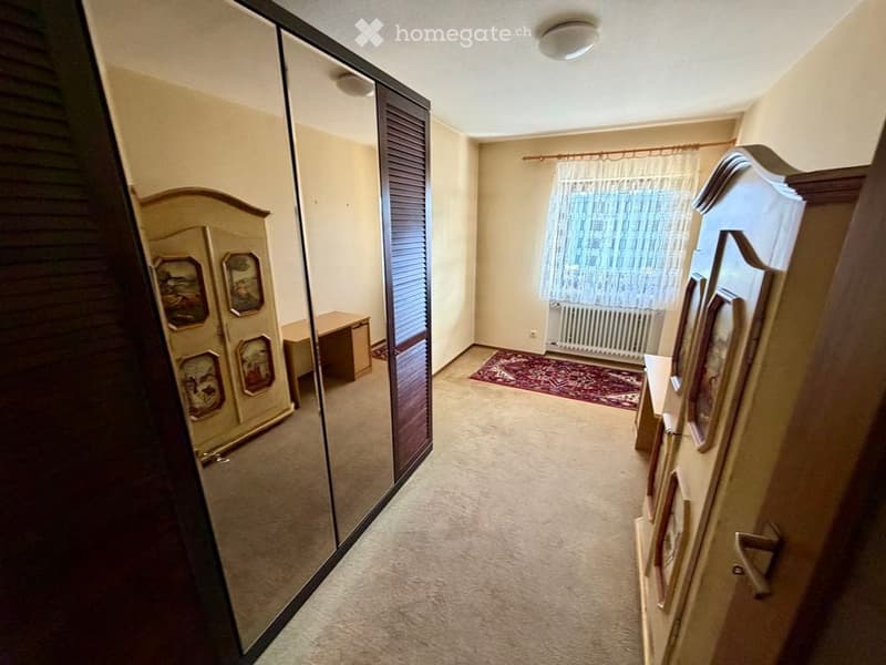 A narrow hallway with carpeted flooring, a large mirrored closet on one side, and a small table with a chair on the other. A window with white curtains and a small red rug are visible at the end of the hallway.