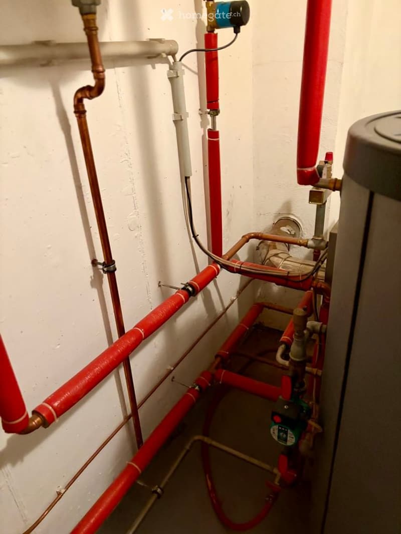 A utility room with exposed red and copper pipes, valves, and a water meter connected to the system.