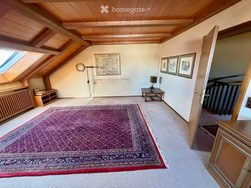 Spacious attic room with wooden ceiling beams, large skylight windows, a patterned area rug, a small wooden table with a lamp, a cabinet, and several picture frames on the walls.