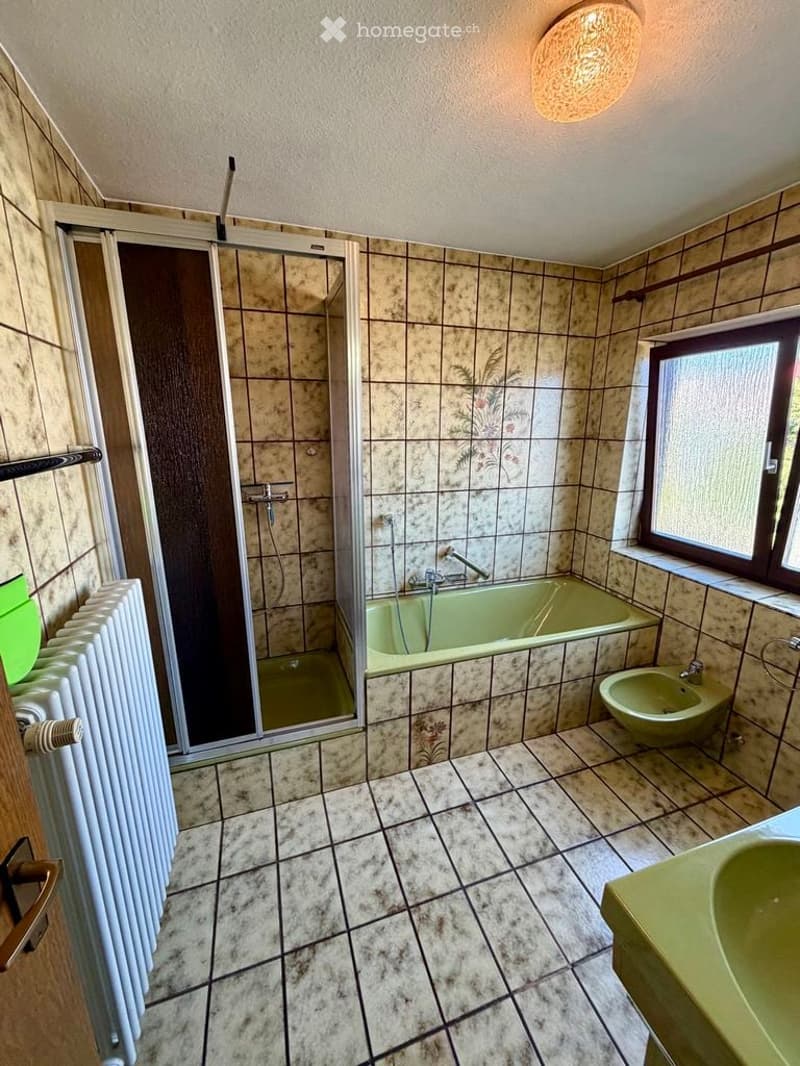 Bathroom with tiled walls and floor, featuring a bathtub, shower, sink, radiator, and bidet. The shower has a glass enclosure, and the room includes a window with frosted glass.