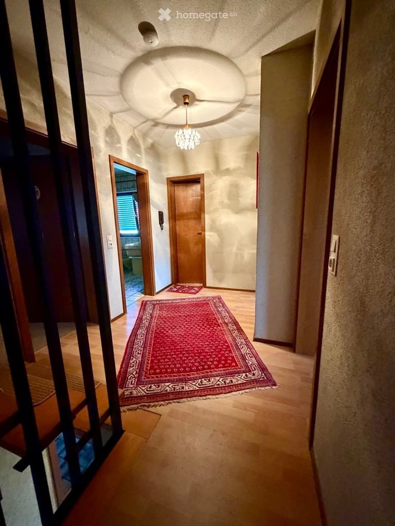 A well-lit hallway with wooden flooring, textured walls, and a decorative red rug. Features include a chandelier, a ceiling light fixture, and several doors leading to other rooms.