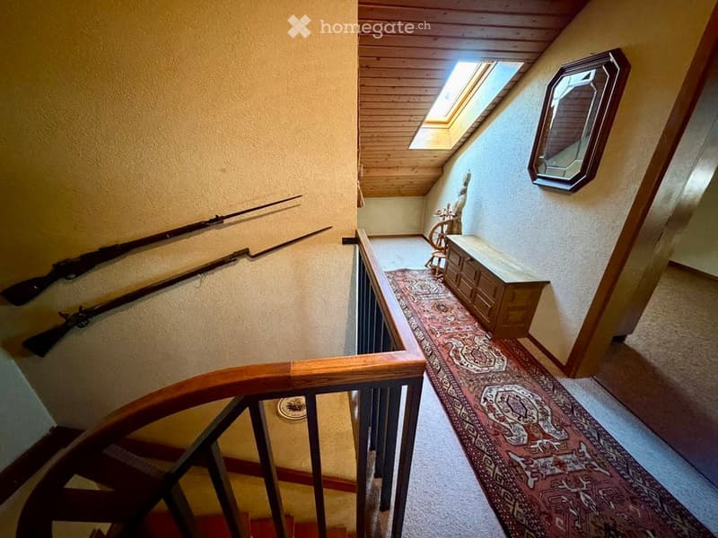 Spacious hallway with wooden ceiling, skylight, patterned carpet, wooden railing, and decorative items including a mirror and a small table.