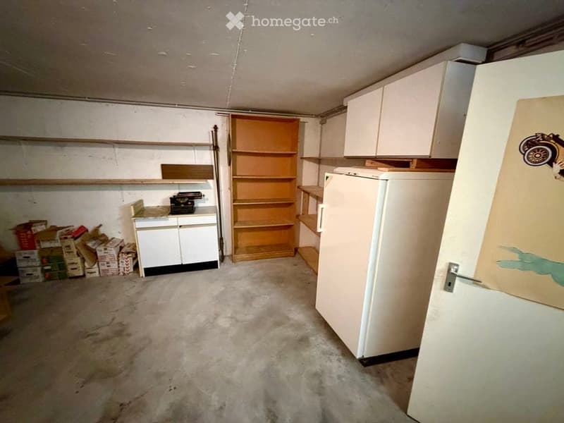 The room features a refrigerator, a wooden shelf, a small table with a typewriter, and several cabinets. The floor is concrete, and there are boxes and storage items scattered around.