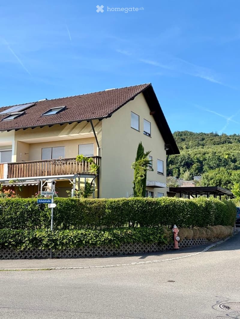 Two-story house with a brown tiled roof, beige walls, and a wooden balcony on the first floor. The house is surrounded by a neatly trimmed hedge and is situated in a residential area with a clear blue sky and green hills in the background.