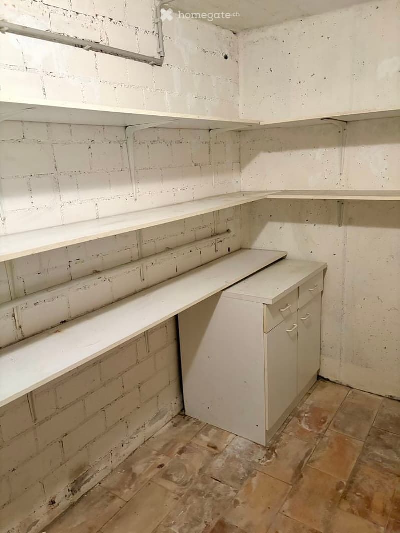 Empty room with exposed brick walls, white shelves, and a small cabinet with drawers. The floor is tiled and appears worn.