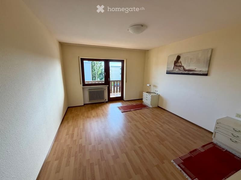 Empty room with wooden flooring, white walls, a window with a door leading to a balcony, a radiator, and a small cabinet with a lamp on top.