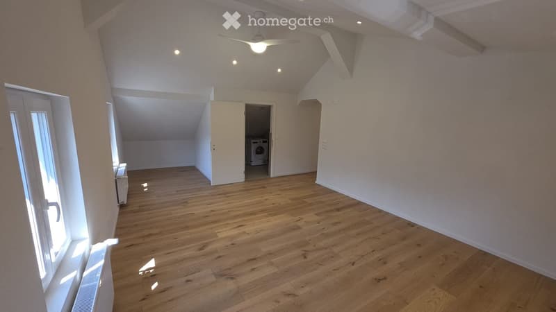 Empty room with wooden flooring, white walls, and a ceiling fan. A doorway leads to another room containing a washing machine.