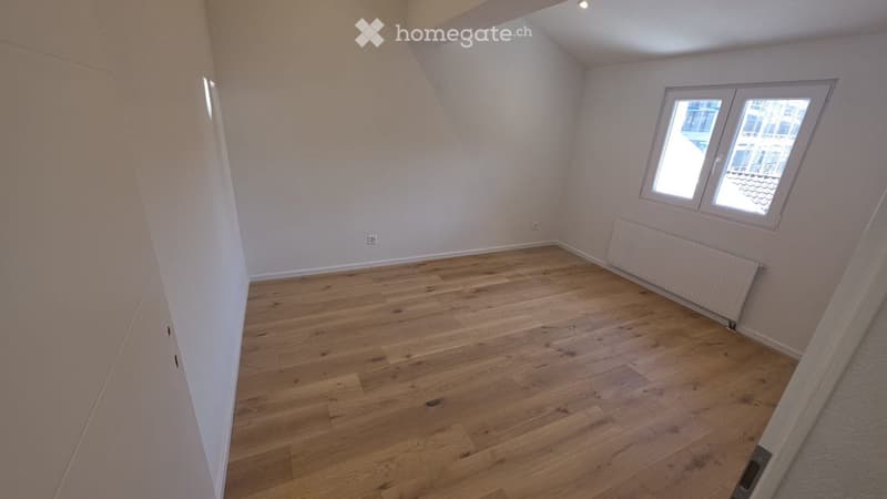 Empty attic room with wooden flooring, white walls, and a single window with white frame. No furniture or equipment present.