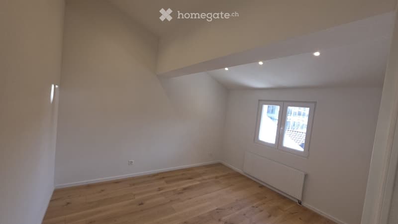 Empty room with wooden flooring, white walls, and a window with a view of the outside. The room features a radiator and recessed ceiling lights.