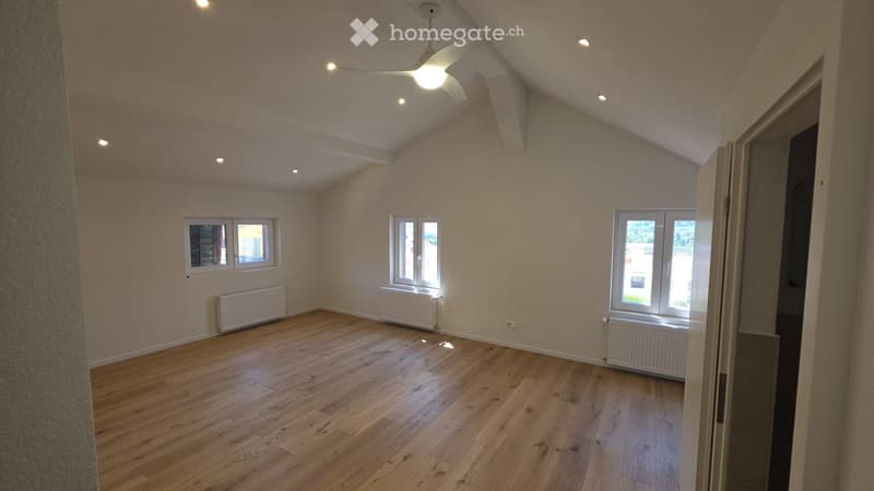 Empty room with wooden flooring, white walls, and multiple windows allowing natural light. Features include a ceiling fan with light, radiators along the walls, and a partially visible adjacent space through an open doorway.