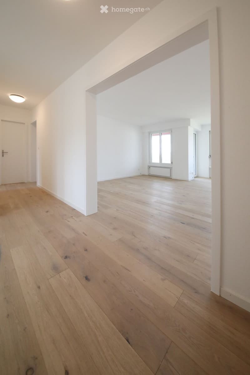 Empty room with light wooden flooring, white walls, and a large window with multiple panes. The room has a simple, modern design with a visible door and a recessed ceiling light.