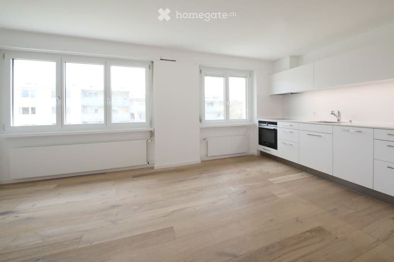 Empty kitchen with white cabinets, wooden flooring, and large windows offering a view of the city.