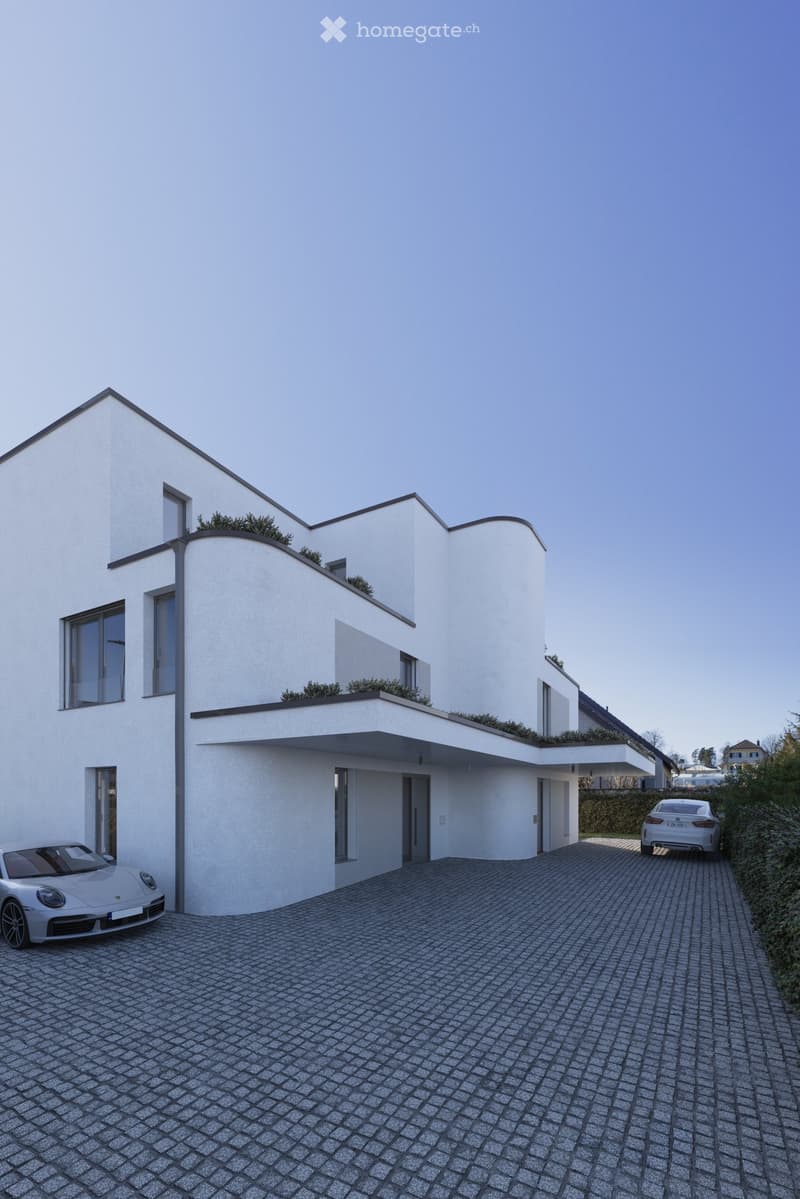 Modern two-story house with white exterior, large windows, and a flat roof with greenery. Two cars parked on a paved driveway.
