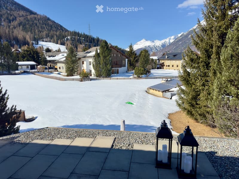 Snow-covered outdoor area with surrounding houses, trees, and mountains in the background. Lanterns with candles are placed on the paved ground.