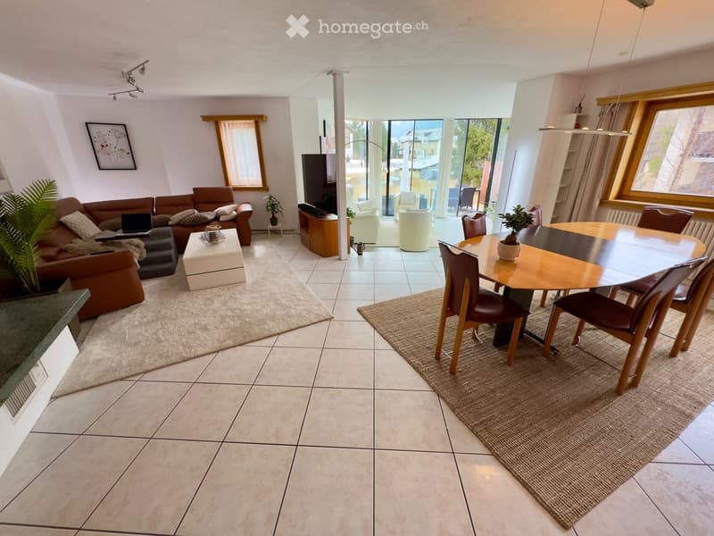 Open-plan living and dining area with tiled flooring, large windows, and a view. Features a brown sofa, coffee table, dining table with chairs, and a television stand.