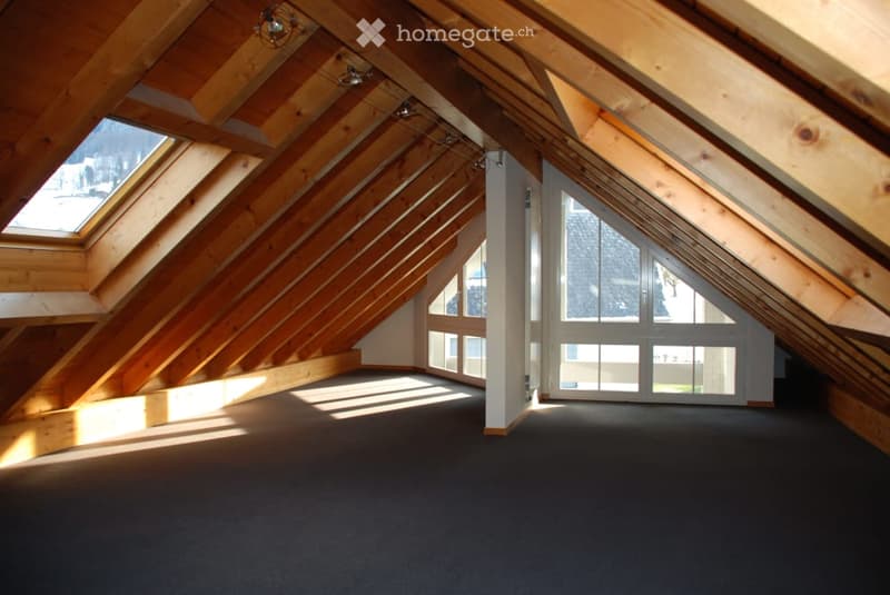 An empty attic space with wooden beams, skylights, and large windows, featuring a dark carpeted floor and exposed wooden ceiling structure.