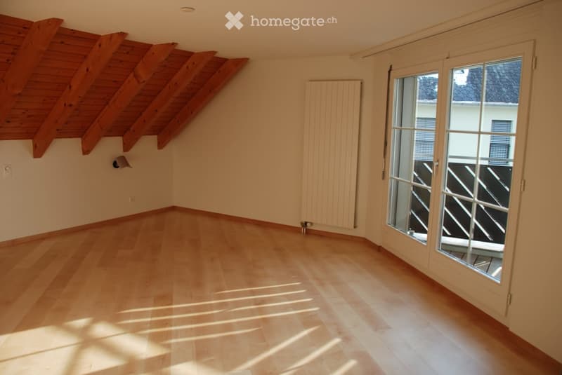 Empty room with wooden flooring, slanted wooden ceiling, large window with glass door leading to a balcony, radiator on the wall, and natural light streaming in.