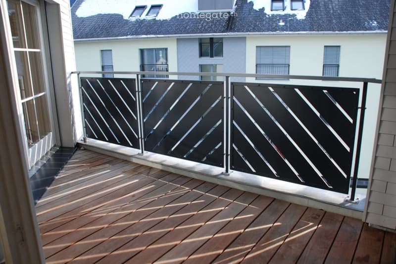 Balcony with wooden decking and modern black and white diagonal patterned railing, overlooking neighboring buildings.