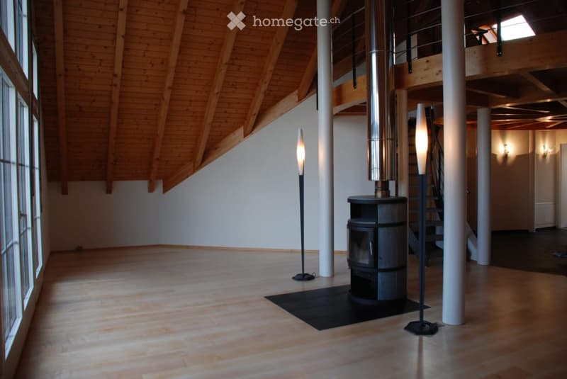 Open, spacious room with wooden floors and exposed wooden beams on the ceiling. A modern black wood-burning stove is centered, flanked by tall, sleek floor lamps. Large windows on one side allow natural light to fill the space.
