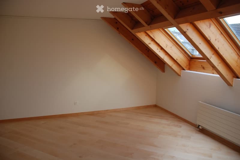 Empty attic room with wooden flooring, white walls, exposed wooden beams, and a skylight window.