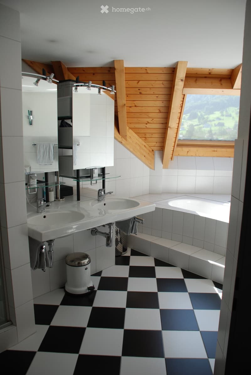 Modern bathroom with double sink, bathtub, checkered floor, wooden slanted ceiling, and large window offering a view.