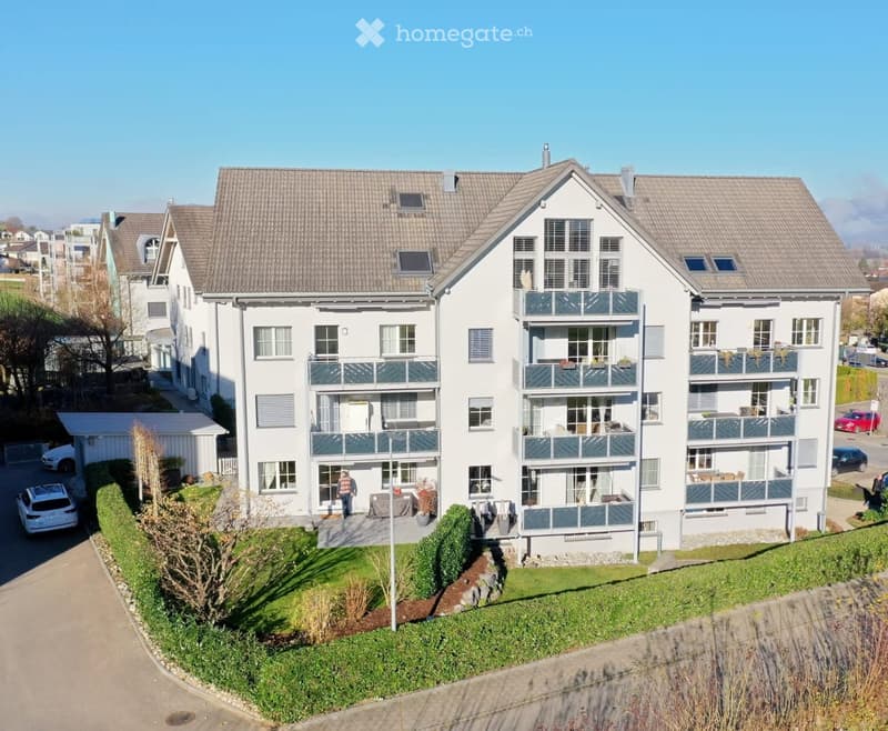 A modern, multi-story residential building with white exterior walls, grey tiled roofs, and multiple balconies. The building features large windows and is surrounded by a well-maintained lawn and driveway with parked cars.
