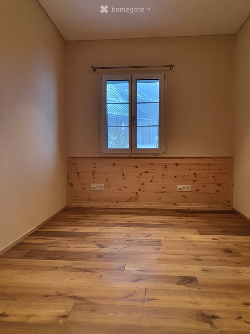 Empty room with wooden flooring, light-colored walls, and a large window with a view of a roof and trees.