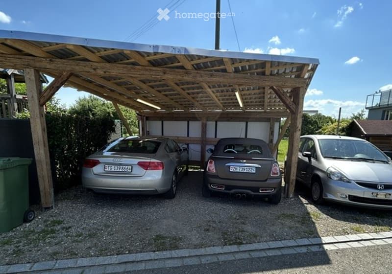 Carport with three parked cars under a wooden roof, gravel ground, and adjacent greenery.