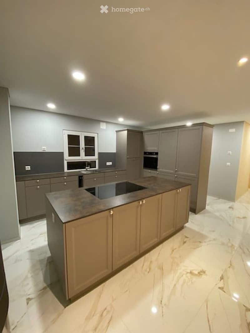 Modern kitchen with a central island, built-in oven, and sleek cabinetry. The space features a marble floor and recessed lighting.