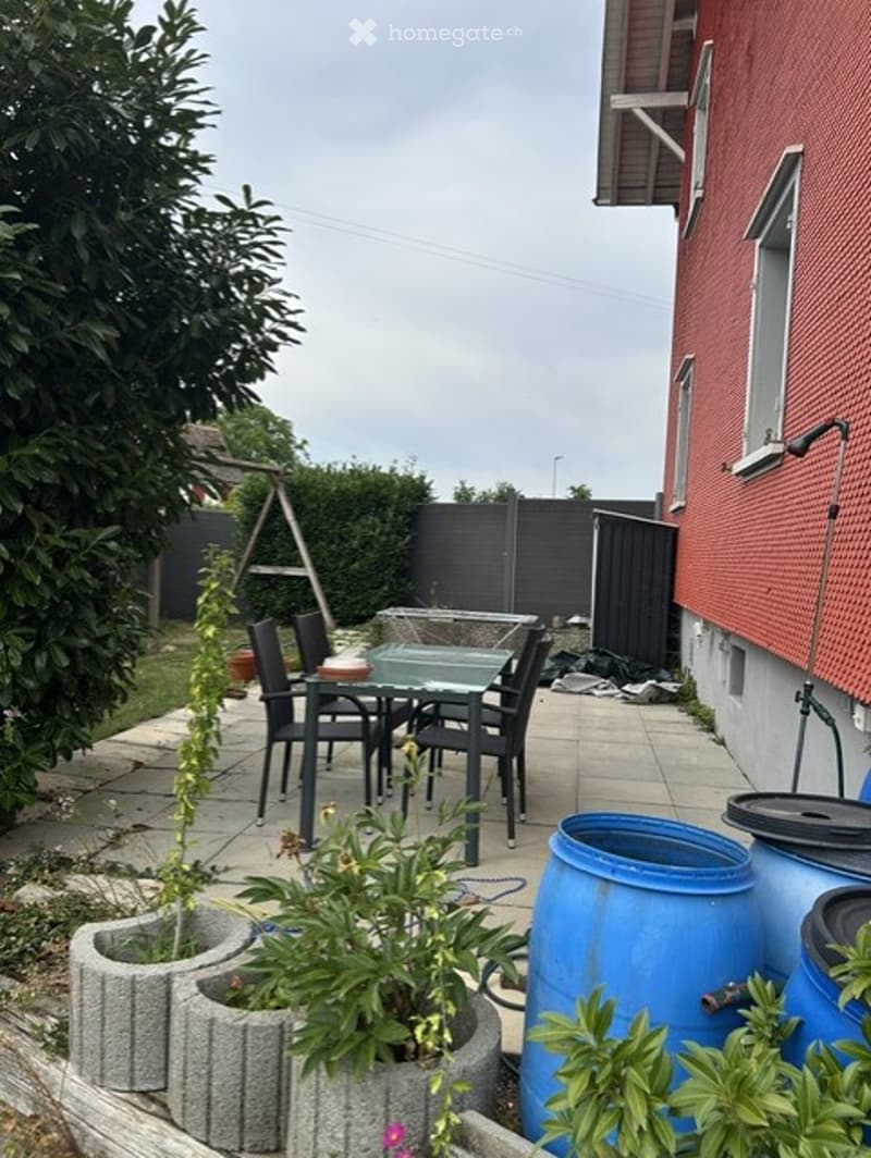 Outdoor patio area with a glass dining table, black chairs, potted plants, and blue barrels. The space is enclosed by a black fence and adjacent to a red brick building.