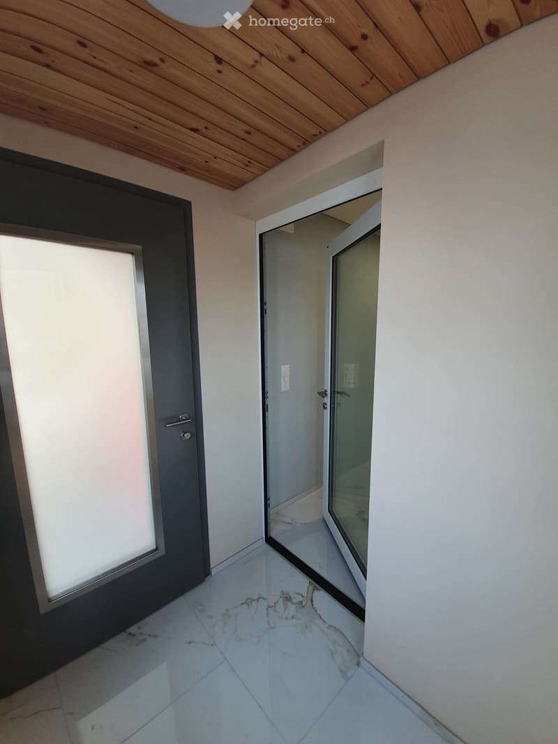 Empty hallway with white walls, marble tiled floor, and wooden ceiling. Features a black door on the left and an open doorway leading to another room on the right.