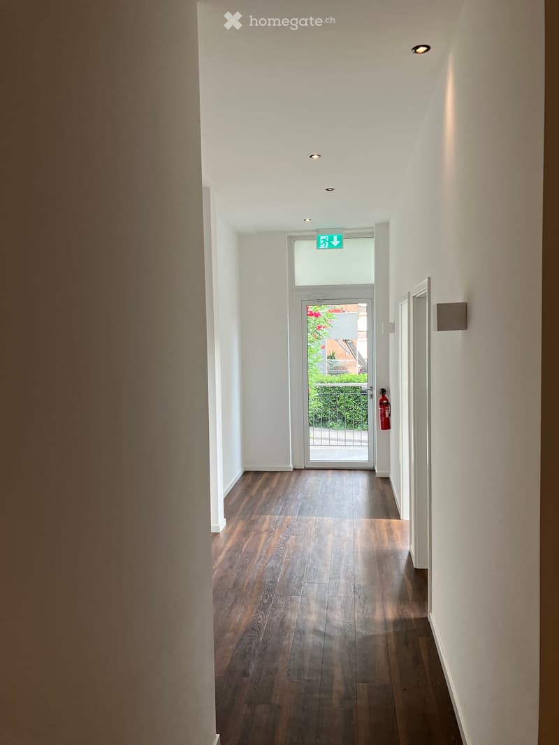 A modern hallway with wooden flooring, white walls, and recessed lighting. An emergency exit sign is visible above a glass door leading outside.