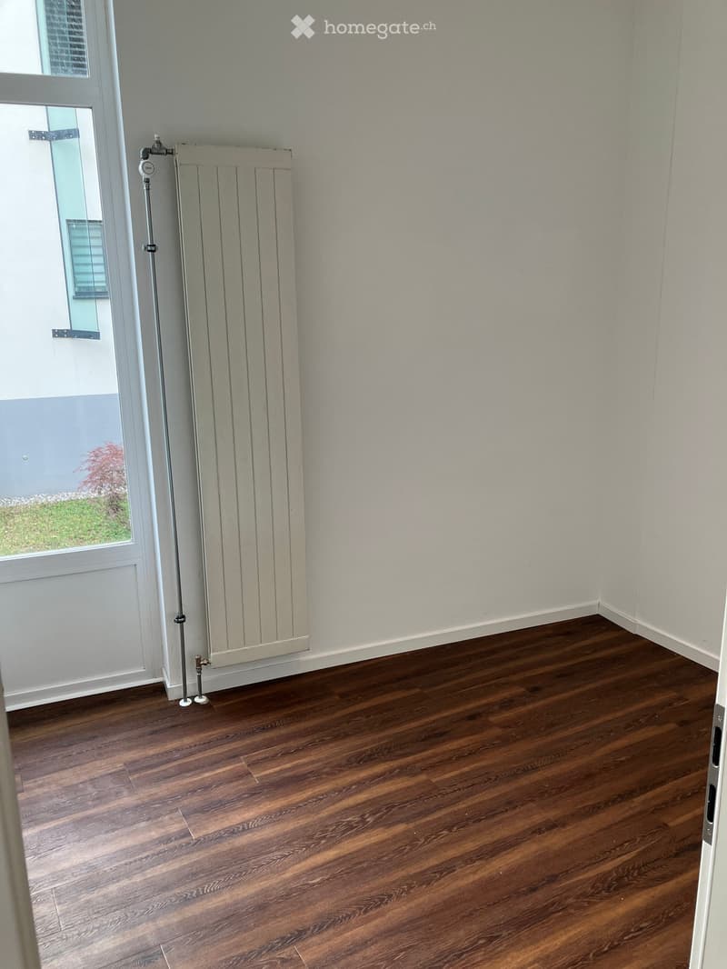 Empty room with wooden flooring, white walls, and a radiator near a window with a view of greenery outside.