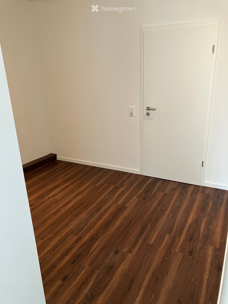 Empty room with wooden flooring and white walls, featuring a closed white door and a light switch on the wall.