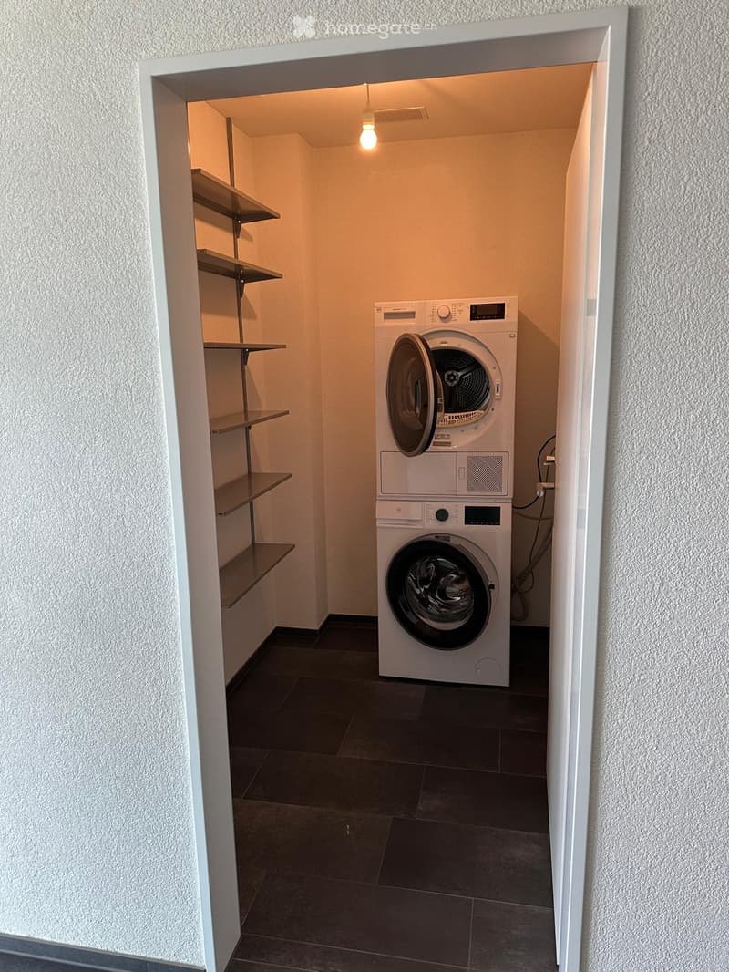 A small, well-lit laundry area with a washing machine, dryer, and metal shelves for storage.