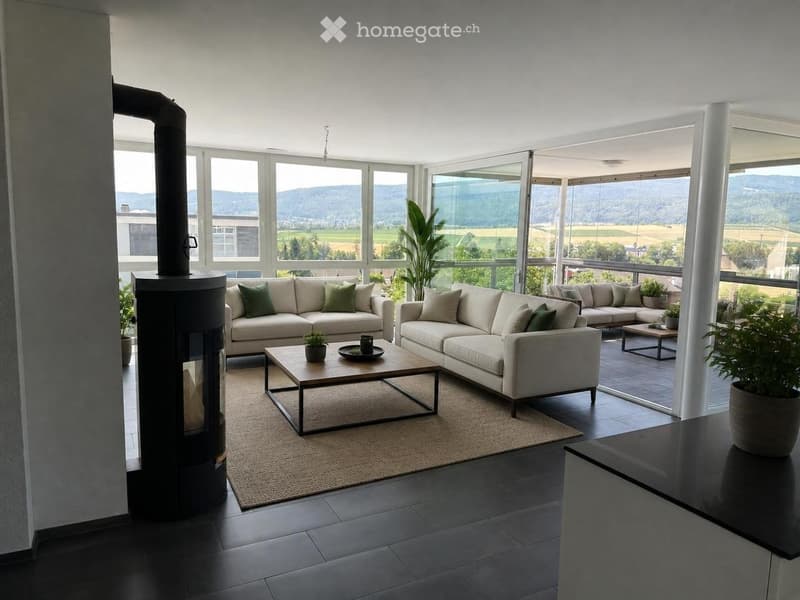 Modern living room with large windows offering a scenic view, featuring a sectional sofa, coffee table, fireplace, and potted plants.