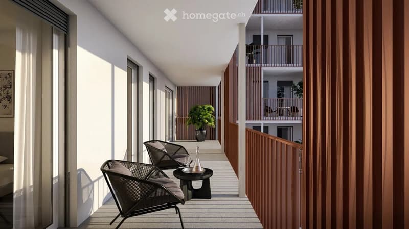 Modern balcony with two wicker chairs, a small round table, and a potted plant. The balcony has wooden railings and is part of a multi-story building with additional balconies visible in the background.