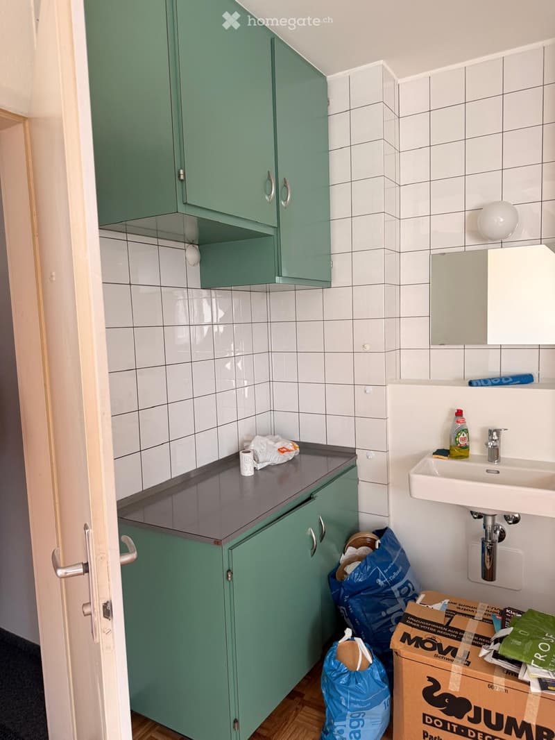 Small bathroom with white tiled walls, green cabinets, a sink, and a mirror. There are some items on the counter and a few boxes and bags on the floor.