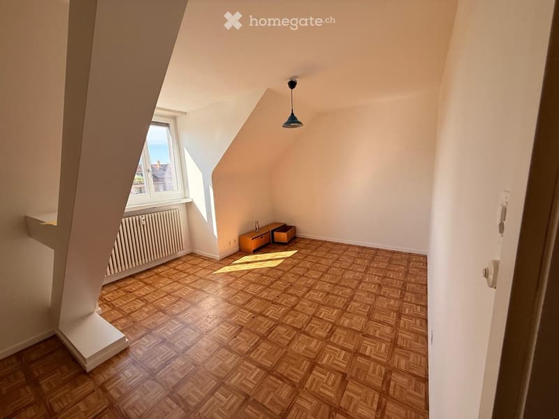 Small, unfurnished room with wooden herringbone flooring, white walls, a window with a view, a radiator, and a small table with storage boxes.