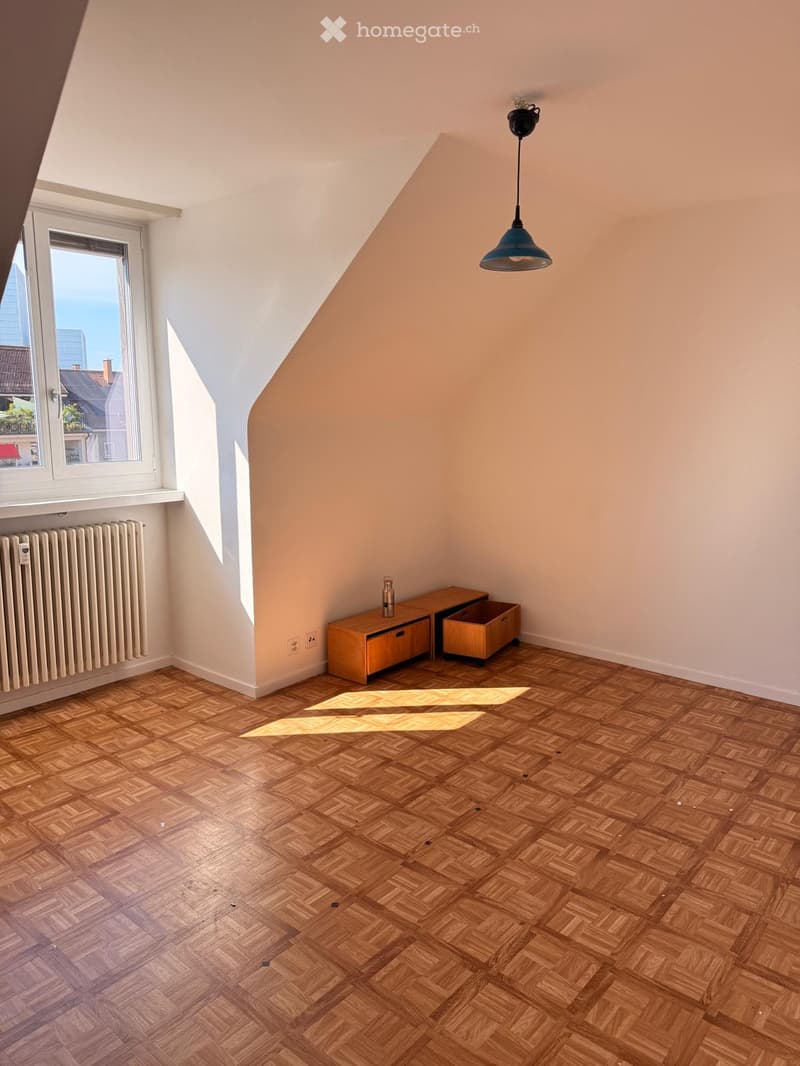 Empty attic room with wooden herringbone flooring, slanted ceiling, large window, radiator, and a small wooden cabinet with drawers.