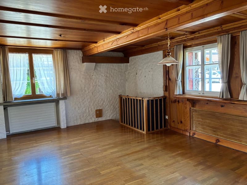 Empty room with wooden floors, exposed wooden beams on the ceiling, large windows with curtains, and a radiator under one window.